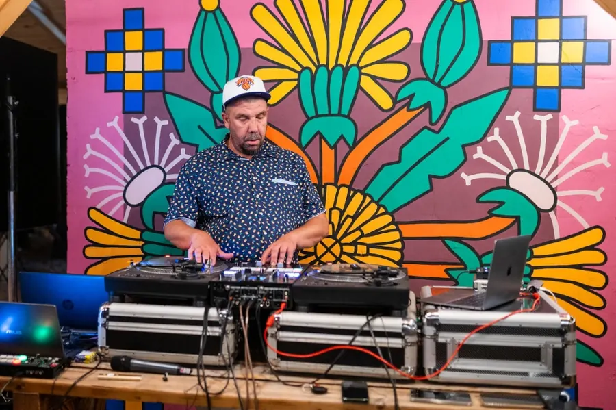DJ za gramofonima na stolu od paleta, ispred oslikanog zida u jarko roze boji.