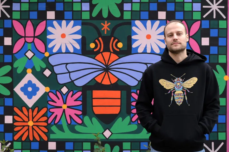 Jedan od braće ispred oslikanog zida sa pčelom i radama, u crnom dukseru sa vezenom pčelom.