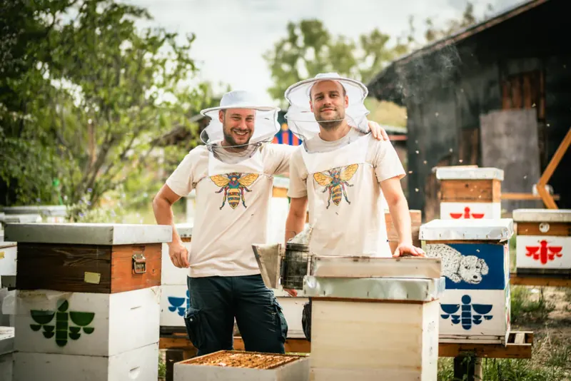 Dario i David Mardešić, velovi zabačeni, ruka preko ramena, među belim košnicama oslikanim znakom pčele.